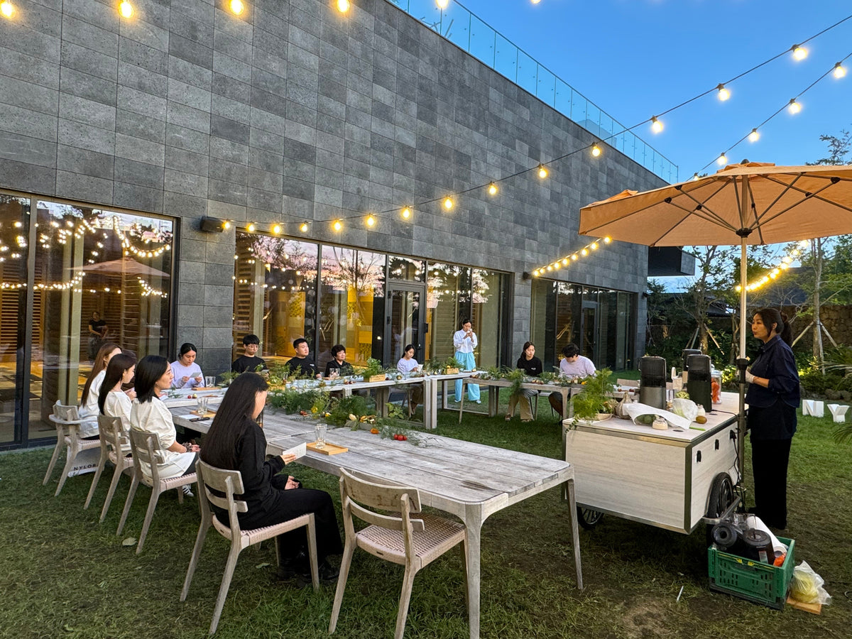 People seated at a long outdoor table in a courtyard with string lights while a person stands nearby